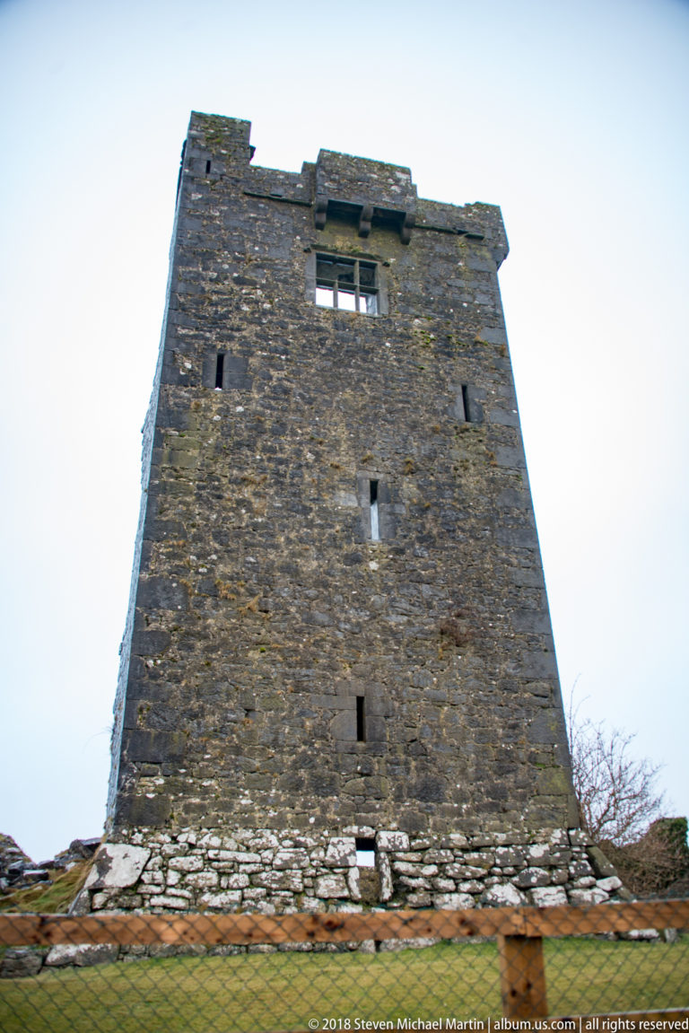 Broad stone face of Dunguaire Castle’s tower house seen from below, with narrow openings, upper window, and parapet.
