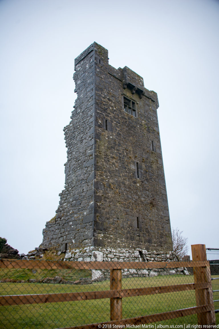 Stone tower house of Dunguaire Castle rises behind a low fence, with narrow windows and weathered masonry visible.