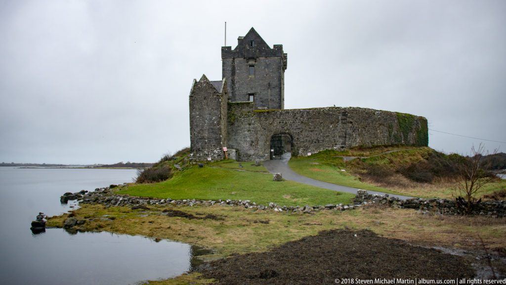 Curving approach road leads to Dunguaire Castle’s arched wall entrance, with the tower house and smaller turret beyond.