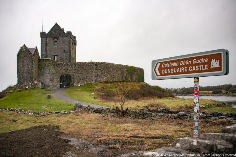 Bilingual Dunguaire Castle directional sign stands beside the approach road, with the stone castle and entrance wall behind it.