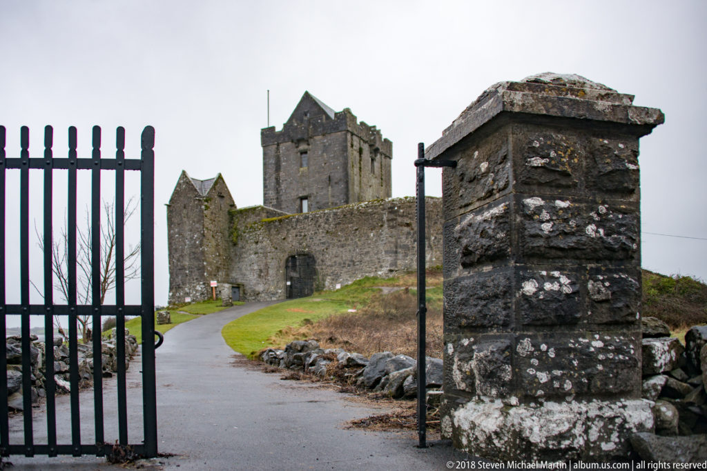Open iron gate and stone pier frame a paved approach to Dunguaire Castle’s wall entrance and tower house.