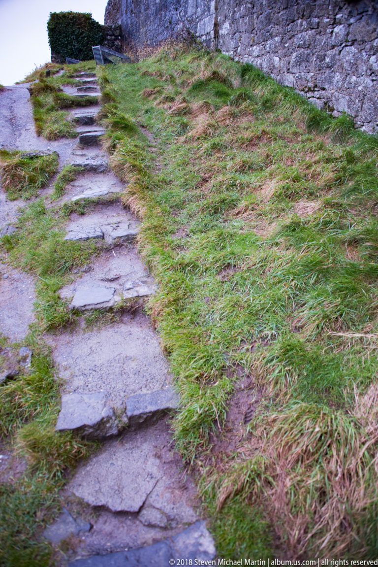 Uneven flat stone footpath crosses wet grass beside a rough stone wall at Dunguaire Castle.