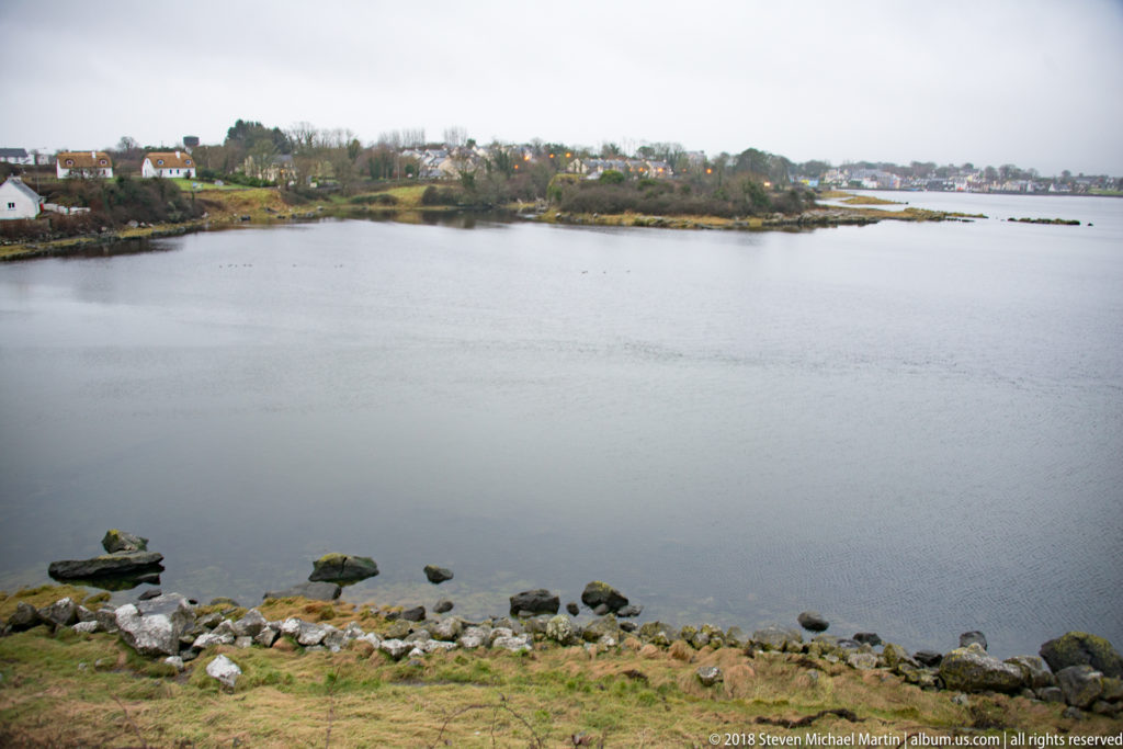 Calm inlet seen from Dunguaire Castle grounds, with shoreline rocks in front and low houses across the water.