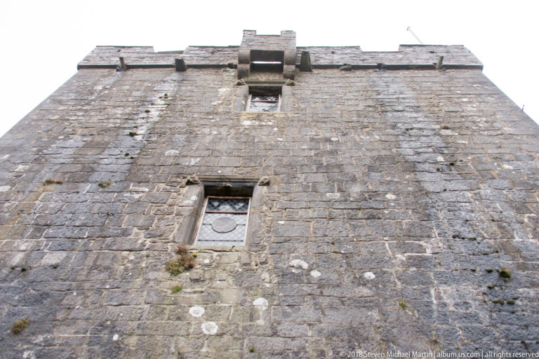 Low-angle view up Dunguaire Castle’s stone tower wall, showing sparse windows, rough masonry, and the parapet above.