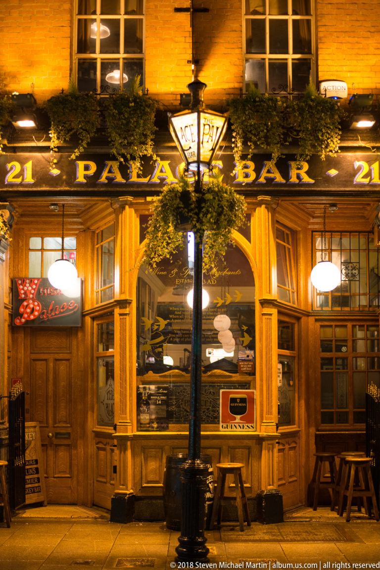 Night view of The Palace Bar’s timber frontage with illuminated signs, hanging baskets, stools, and a streetlamp.