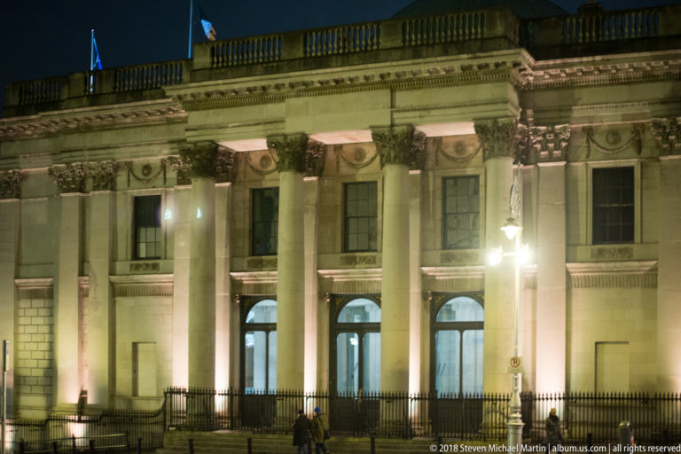 Illuminated neoclassical façade with tall columns, carved capitals, recessed doors, upper balustrade, and iron railings.