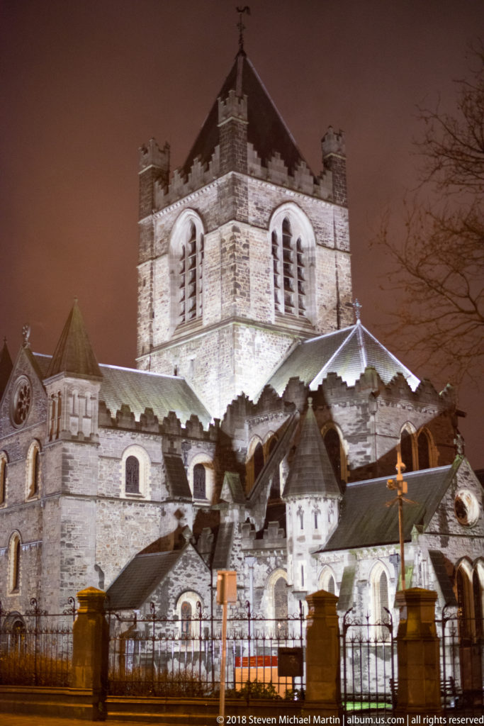 Floodlit Christ Church Cathedral at night with square central tower, layered roofs, stone walls, and perimeter railings.