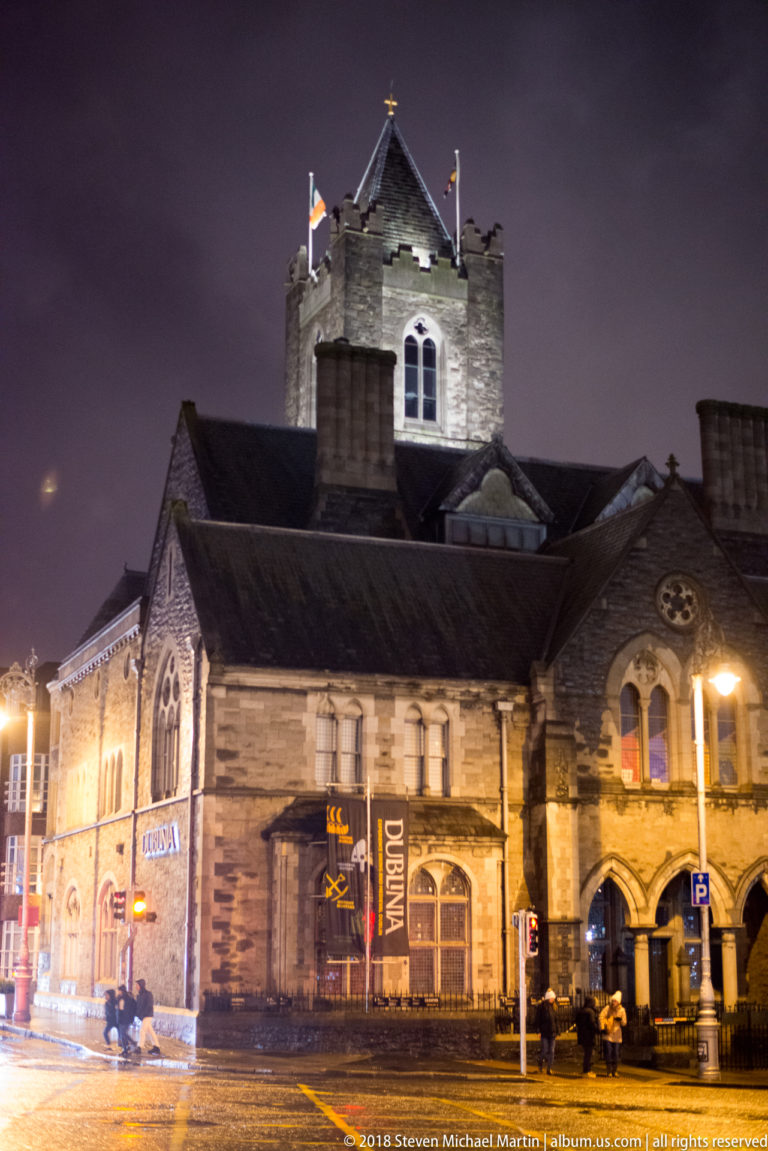 Night street view of the Christ Church Cathedral precinct with the cathedral tower rising behind lower historic buildings.