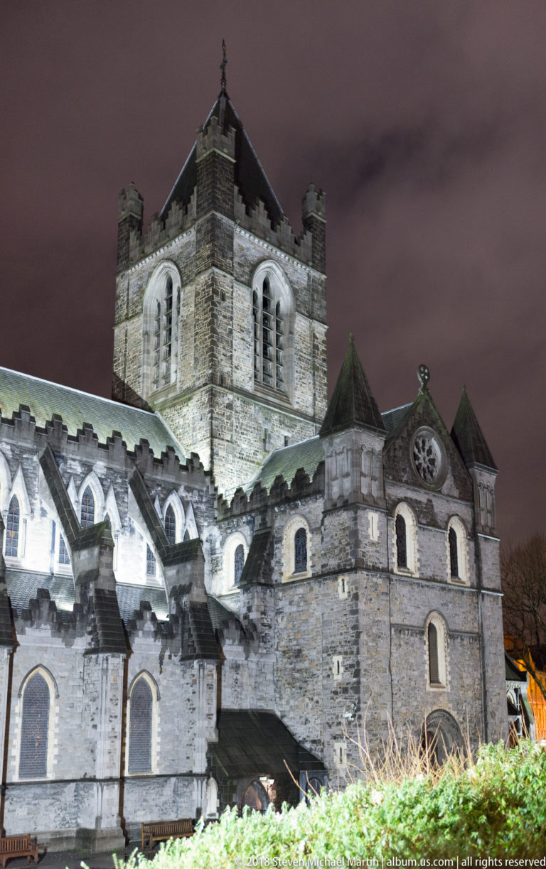 Side view of floodlit Christ Church Cathedral with square tower, buttressed walls, arched windows, and layered roofs.