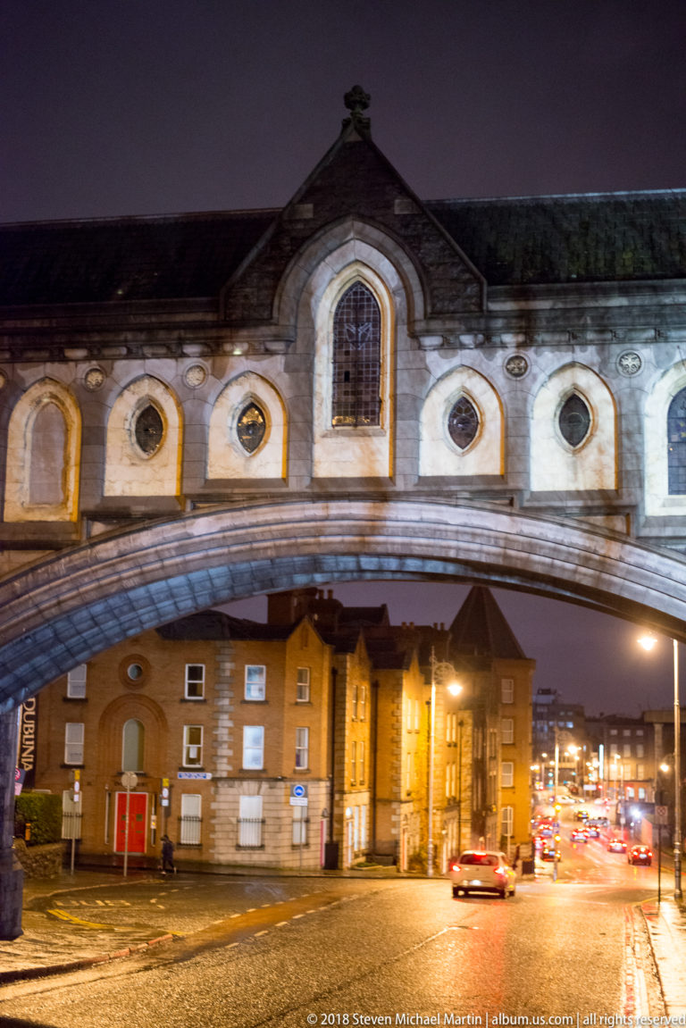 Street passes beneath the enclosed Christ Church–Dublinia bridge, with Gothic-style windows above and traffic beyond.