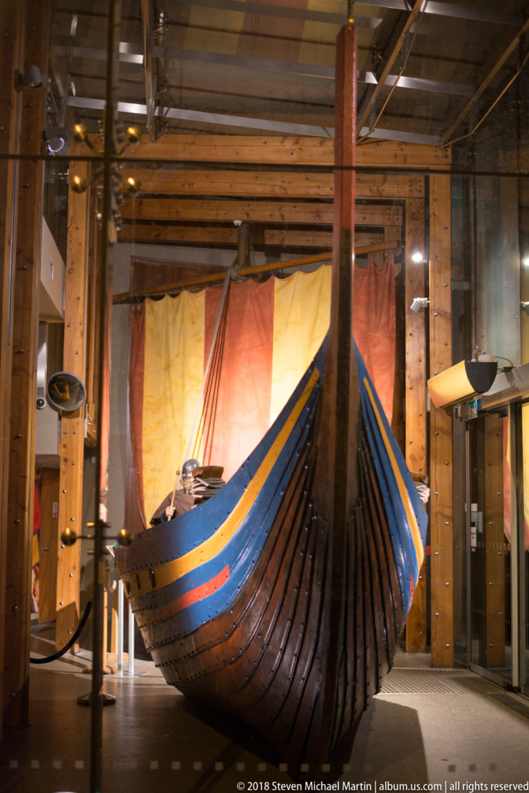 Indoor museum display of a clinker-built Viking-style wooden boat with high curved ends and a striped sail behind it.