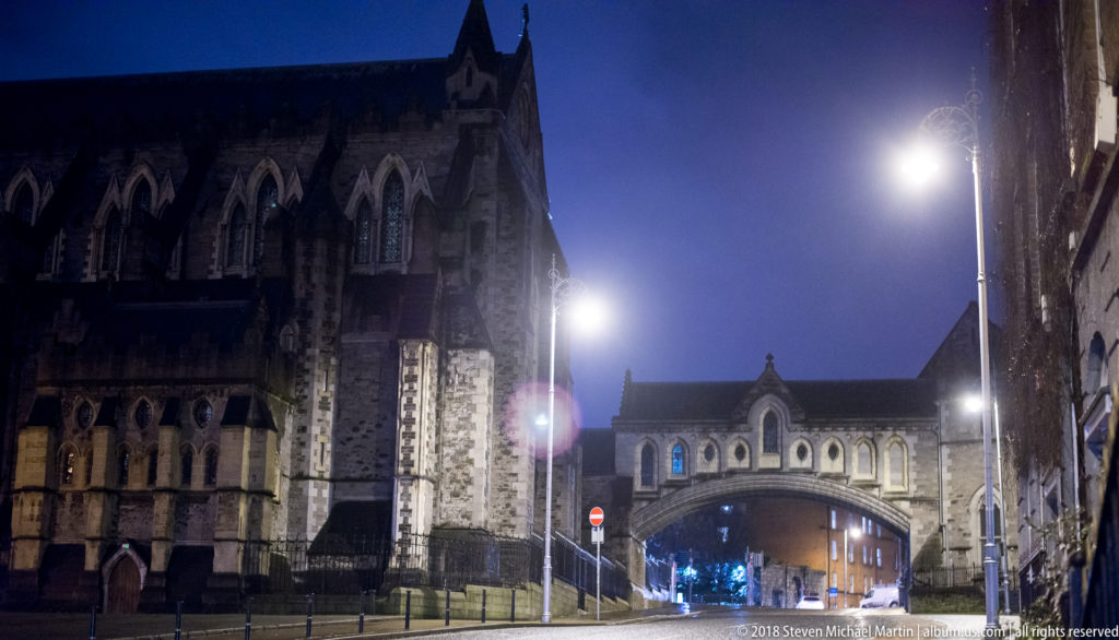 Wide night street view with Christ Church Cathedral at left and the enclosed Dublinia bridge spanning the road ahead.