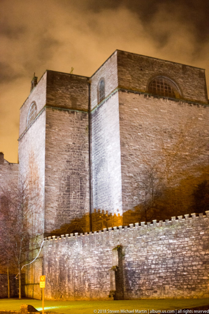 Floodlit massive stone walls behind a lower crenellated wall in the Dublin Castle precinct at night.