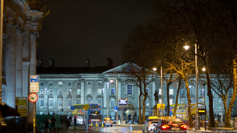 Wet Dublin street at night facing a floodlit neoclassical building, with buses, cars, trees, and streetlamps.