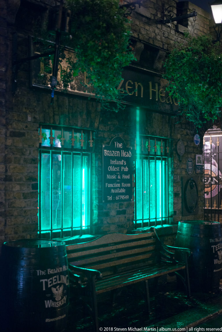 Night entrance to The Brazen Head pub with green-blue lighting, signs, benches, plaques, and hanging plants.