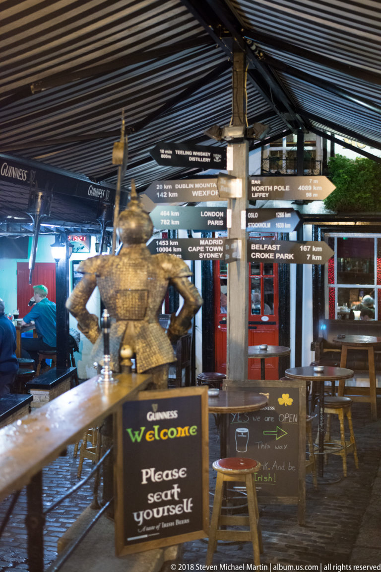 Decorative armored figure in The Brazen Head courtyard beside a welcome sign, directional boards, and pub tables.