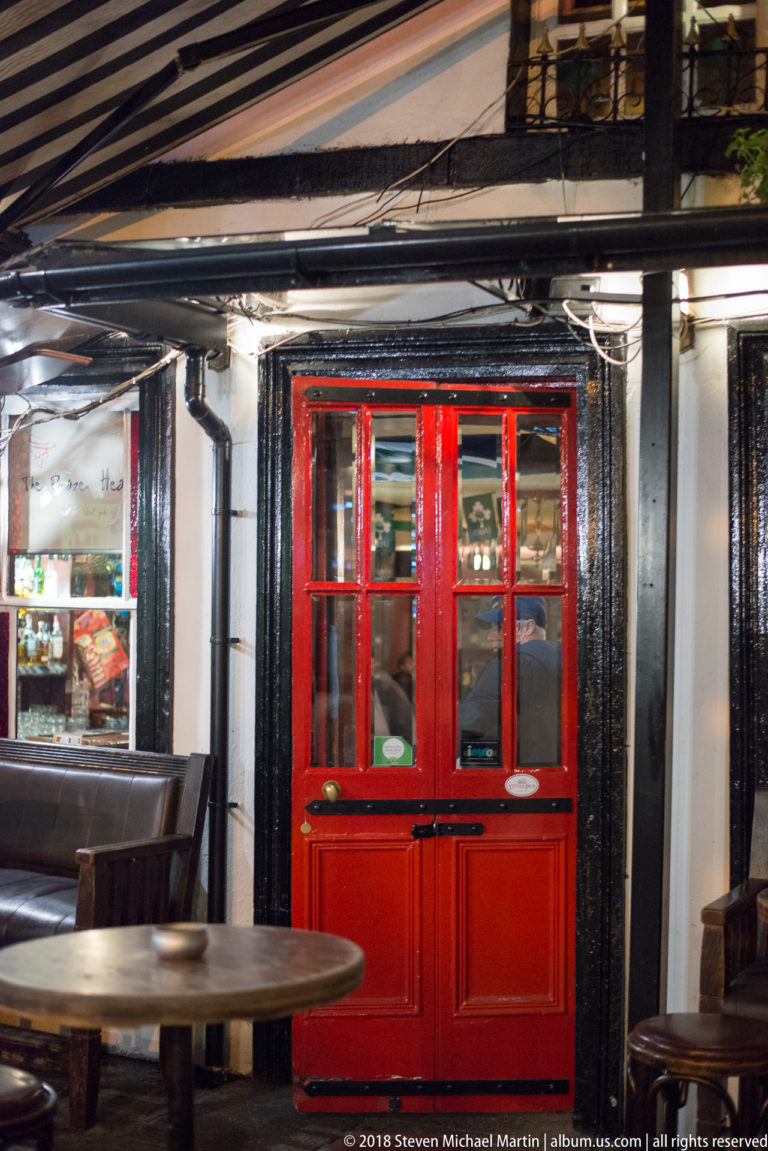 Red glazed pub door set in glossy black timber paneling, with mirrors and warm interior reflections around it.