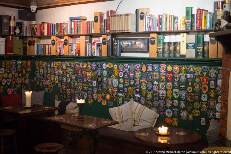 Pub seating nook with a wall densely covered in colorful patches, books above, candles, tables, and cushioned benches.