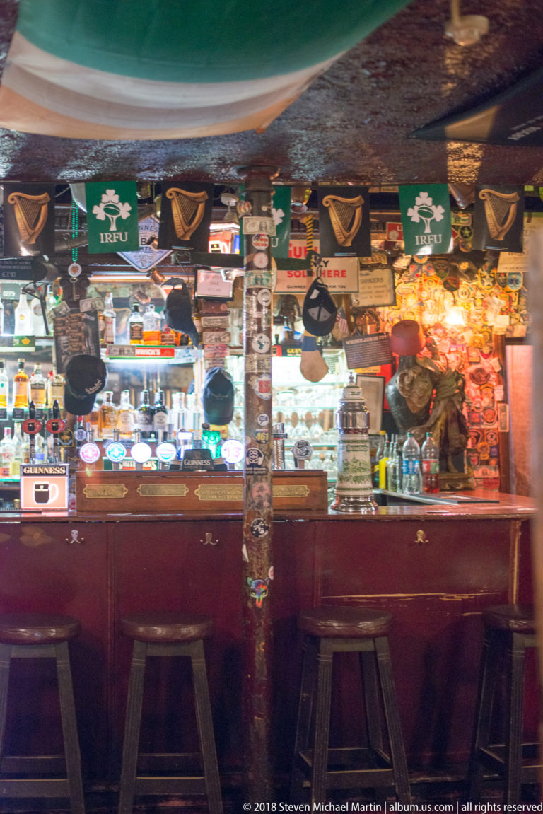 Traditional Dublin pub bar with three stools, bottles, Irish-themed flags, signs, hats, and dense memorabilia.
