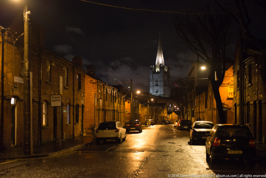 Wet Dublin residential street at night with brick terraced houses, parked cars, and a brightly lit church spire ahead.