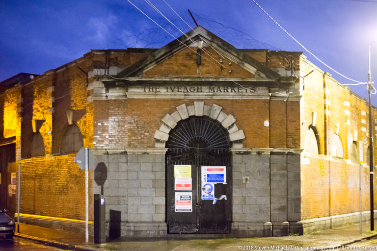 Night view of the Iveagh Markets entrance with pedimented sign, large arched gateway, brick walls, and closed metal gates.