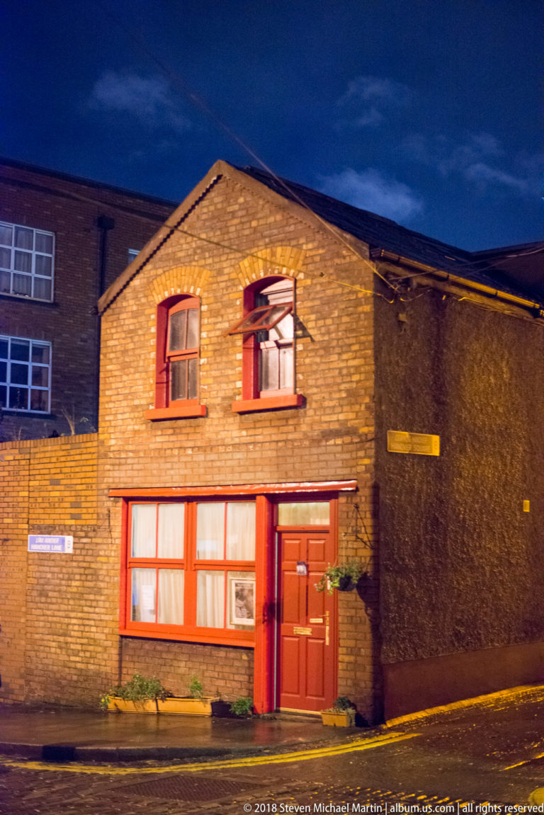 Two-storey brick house at night with red-painted door and window trim, gabled roof, and minimal street setback.