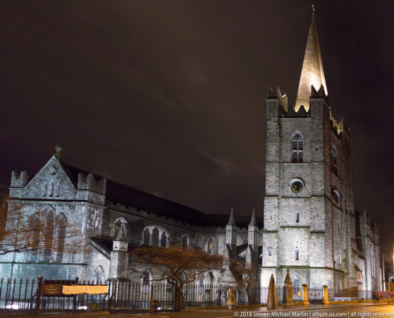 St Patrick’s Cathedral at night with tall square tower, lit spire, Gothic walls, trees, and perimeter railings.
