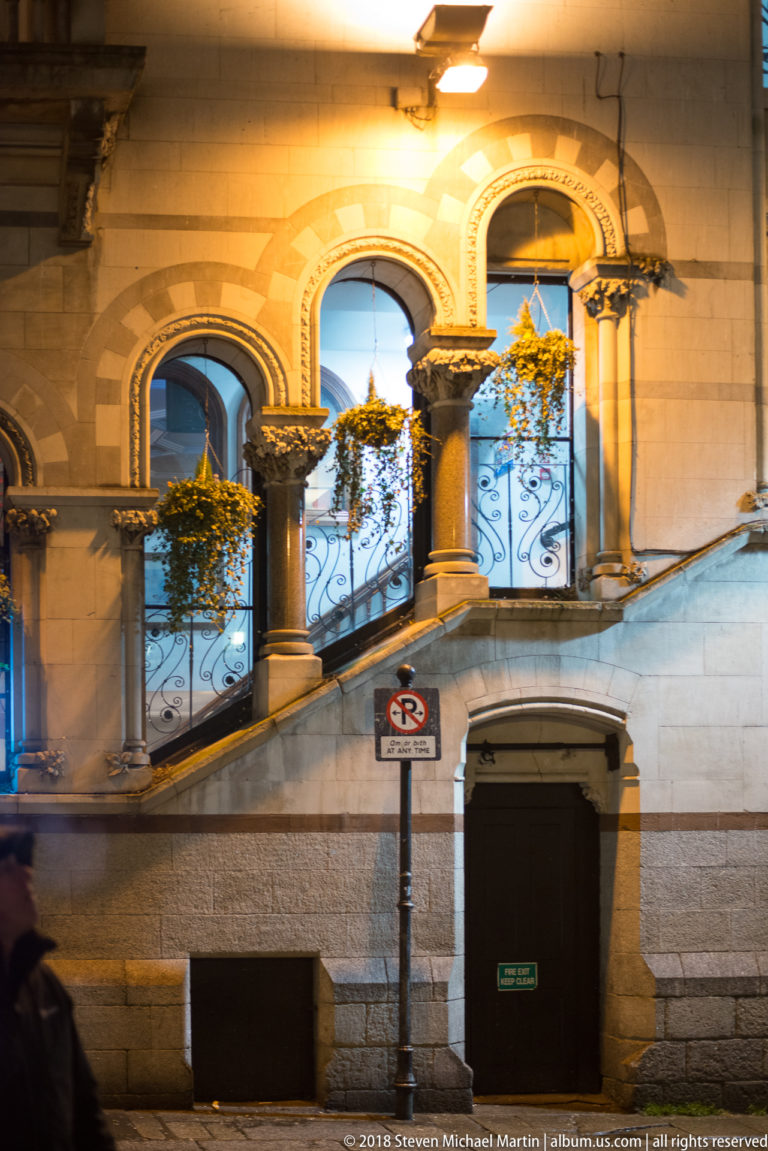 Illuminated historic stone façade with three arched windows, iron balcony railings, hanging baskets, and carved ornament.
