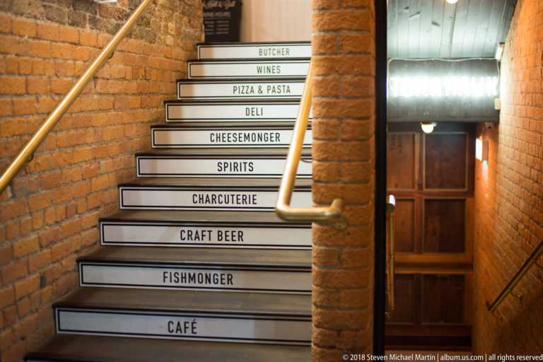 Brick stairwell with illuminated step signs listing café, fishmonger, craft beer, charcuterie, spirits, deli, wines, and butcher.
