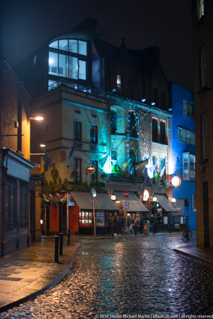 Wet cobblestone street at night with blue-lit façades, pub fronts, and an overhead sculptural installation.