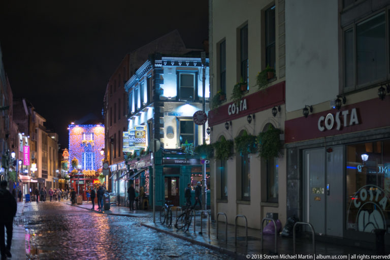 Narrow Dublin street at night with pedestrians, bicycles, pub façades, wet paving, and bright decorative lights in the distance.