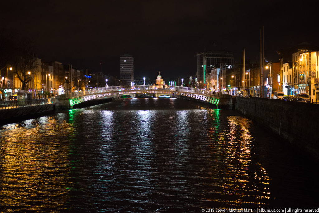 River Liffey at night with an illuminated pedestrian bridge, quayside lights, and long reflections on dark water.