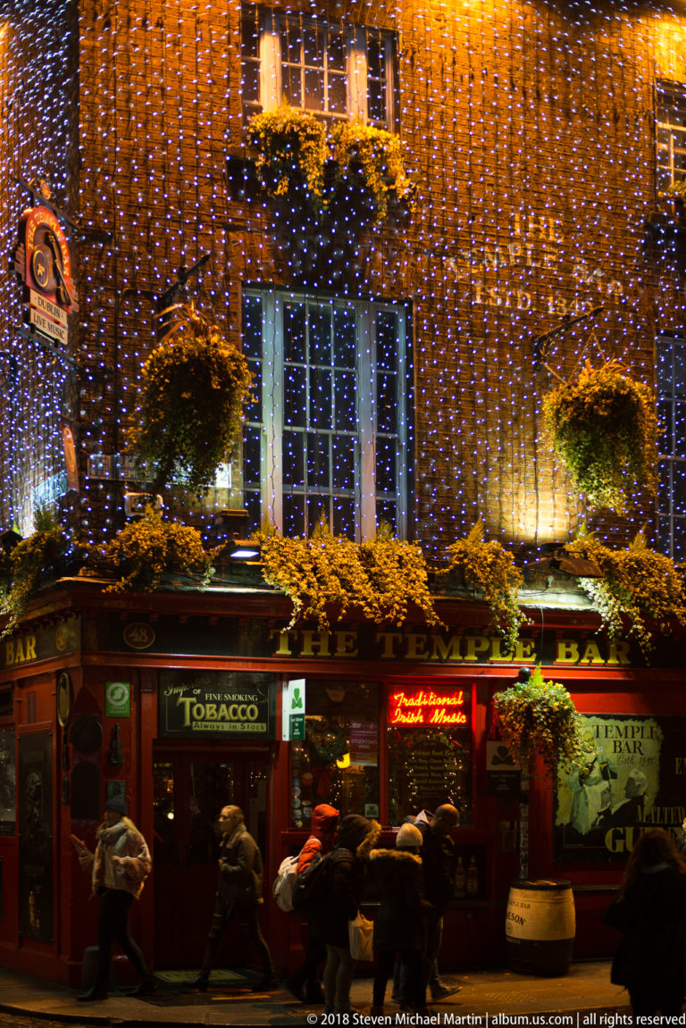 Dublin pub exterior at night covered in orange and blue lights, with hanging baskets, signs, and patrons at the entrance.