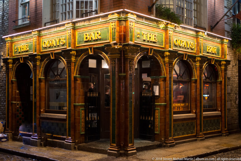 The Quays Bar at night with gold lettering, arched openings, dark glazed panels, and ornate illuminated woodwork.