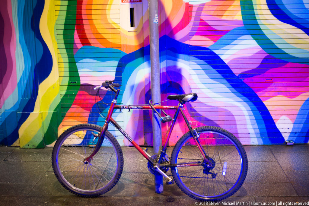 Red bicycle parked against a brightly colored abstract mural of sweeping pink, blue, green, yellow, and violet bands.