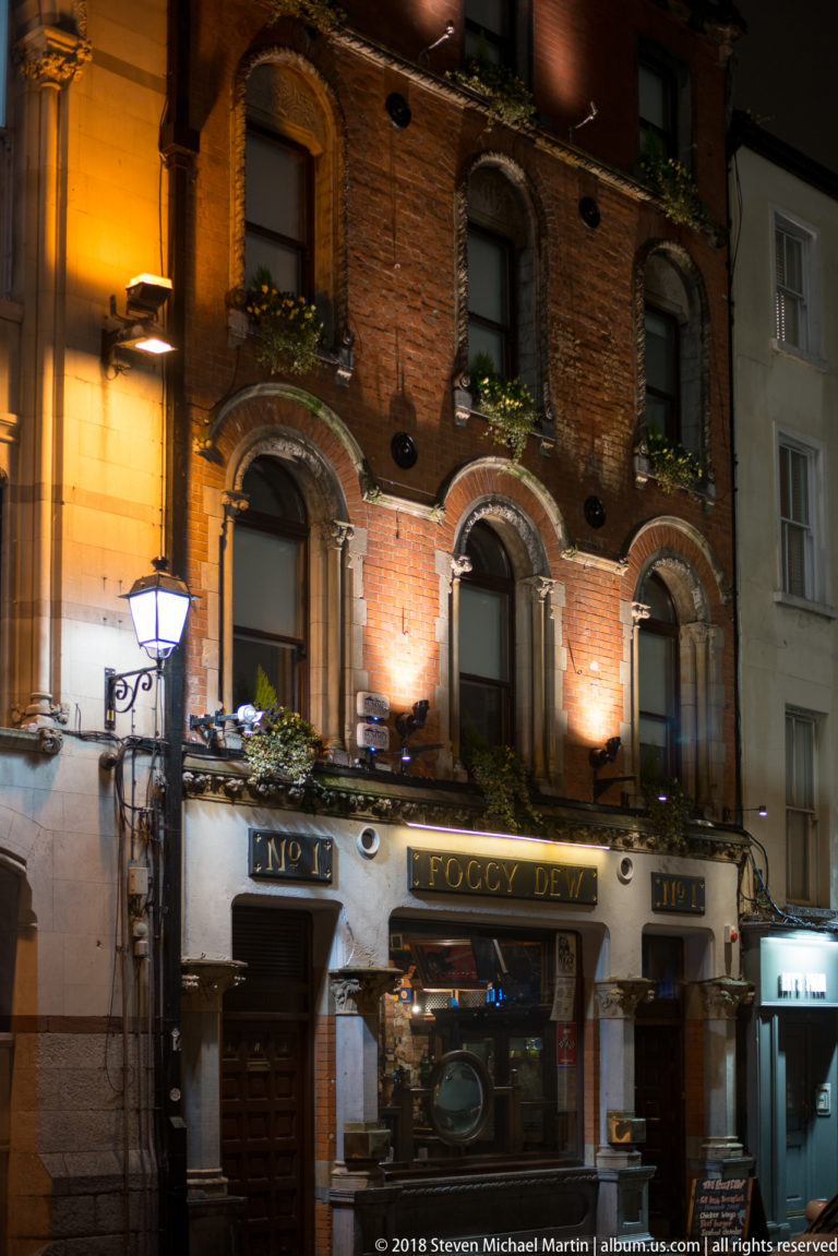Brick pub façade at night with arched windows, flower boxes, carved stone trim, and an ornate ground-floor entrance.