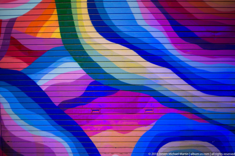 Close detail of an abstract mural with sweeping blue, violet, green, pink, and peach bands across a paneled wall.