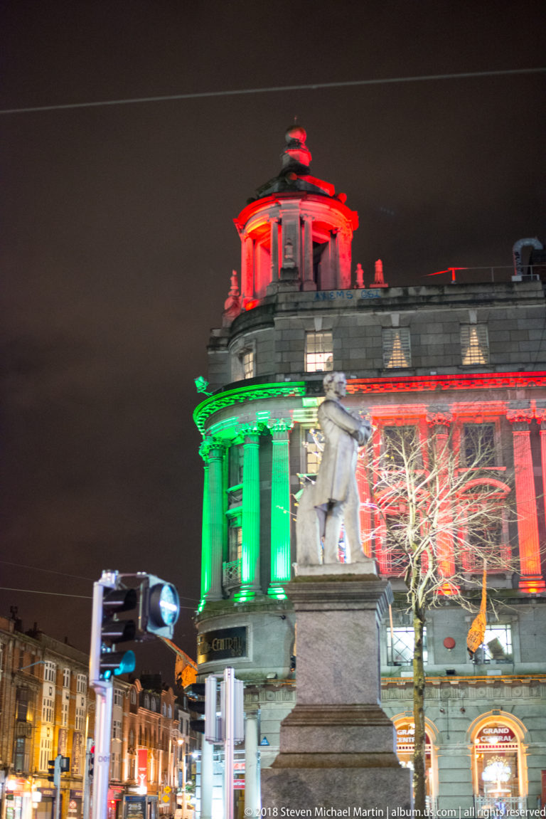 Stone monument before a large Dublin building illuminated in green and red, with street signs and vehicles below.