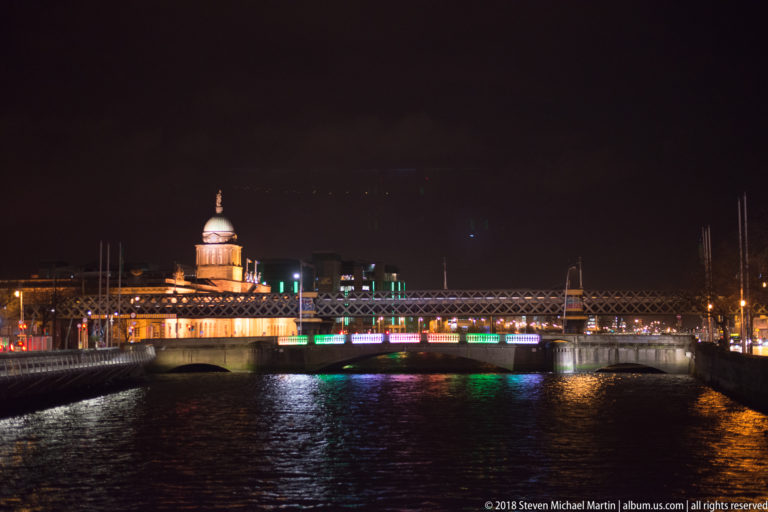 The Custom House illuminated beyond a bridge over the River Liffey, with green and white reflections on the water.