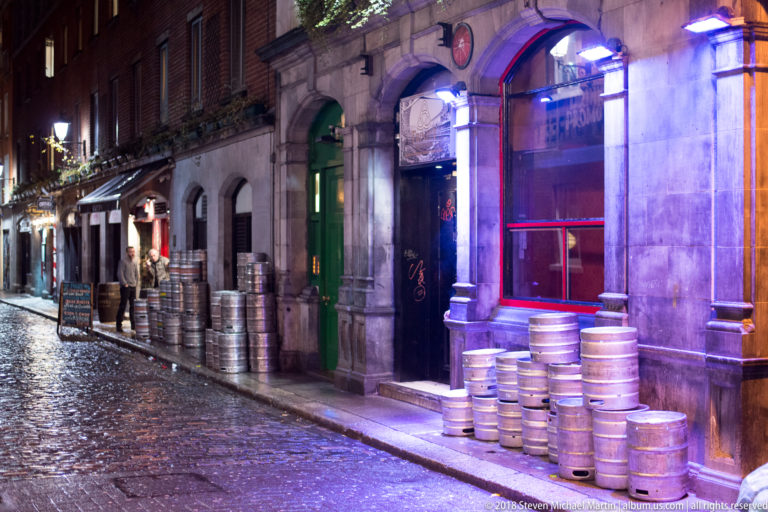 Narrow wet cobbled lane at night with stacked beer kegs outside brightly lit pub entrances.