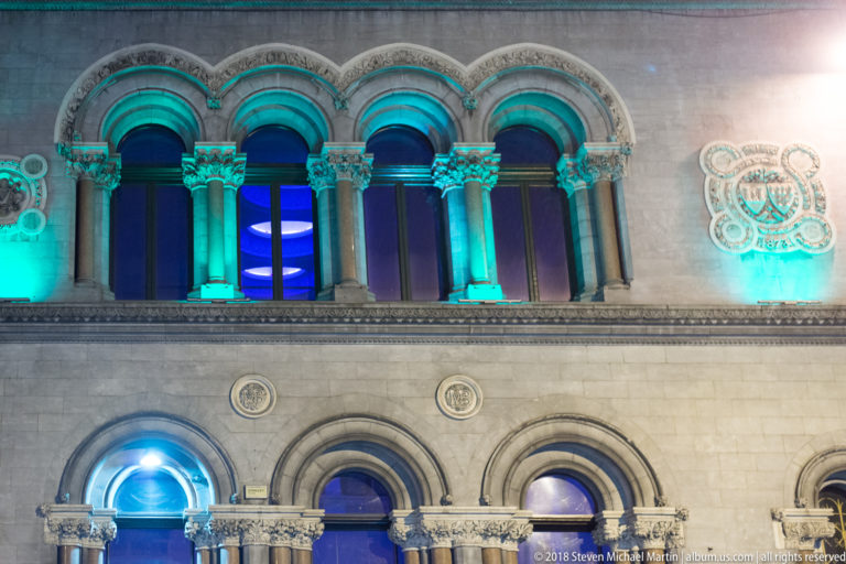 Night detail of a stone façade with round-arched windows, carved columns, capitals, and interlaced circular ornament.