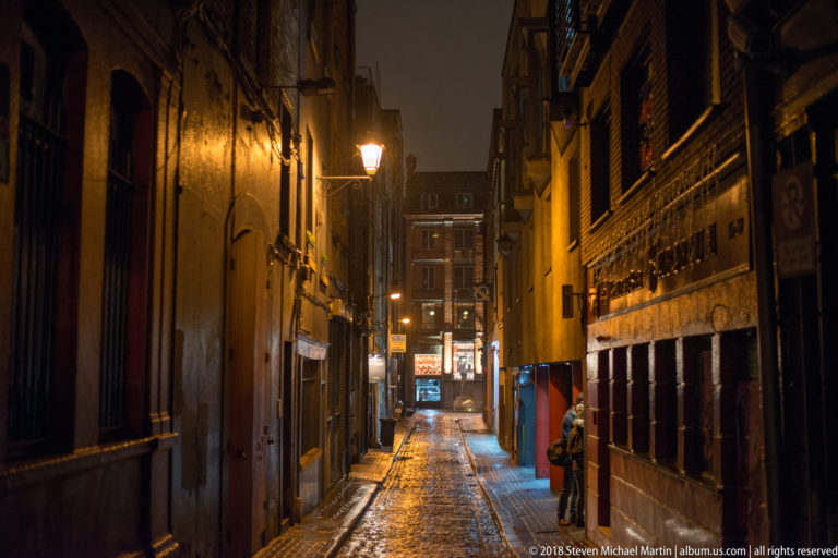 Narrow wet Dublin alley at night, lined with masonry buildings, shopfronts, and traditional streetlamps.