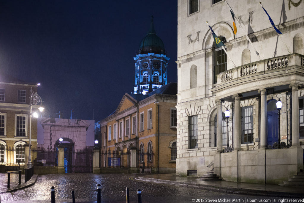 Night view of a paved Dublin civic space with historic buildings, flags, a tower, bollards, and a monument.