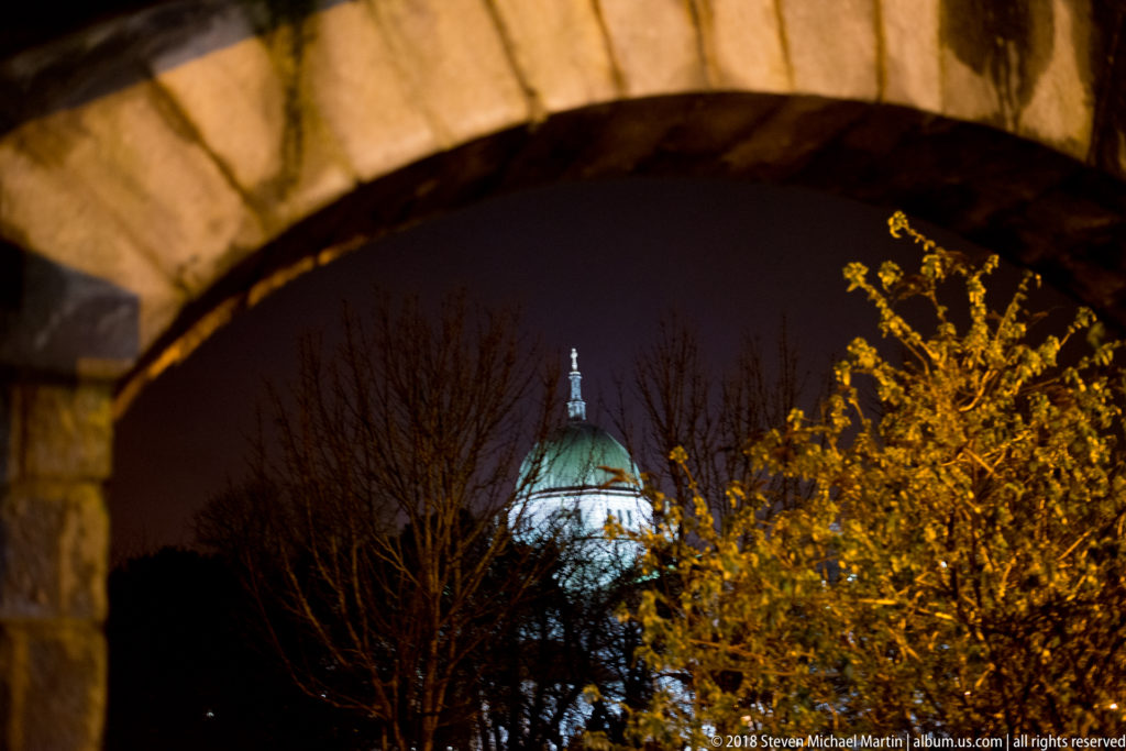 Galway Cathedral's green dome seen through a large stone arch, with branches and yellow leaves below.