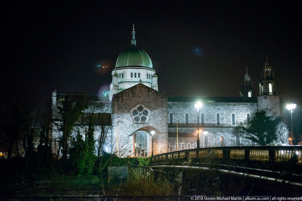 Illuminated Galway Cathedral with green dome and stone façade beyond bridge and riverside railings at night.