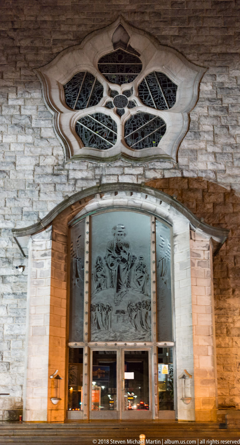 Galway Cathedral entrance with glass doors, sculptural panels, pale stone surround, and multi-lobed rose window above.