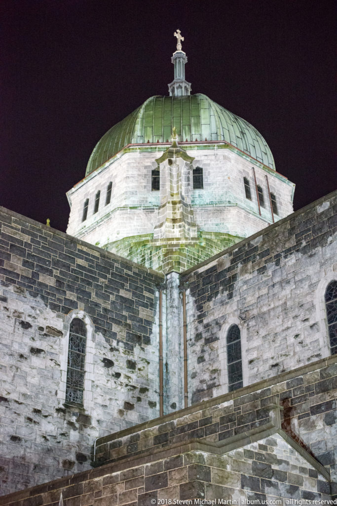 Upward night view of Galway Cathedral's green dome, lantern, cross, and pale stone walls.
