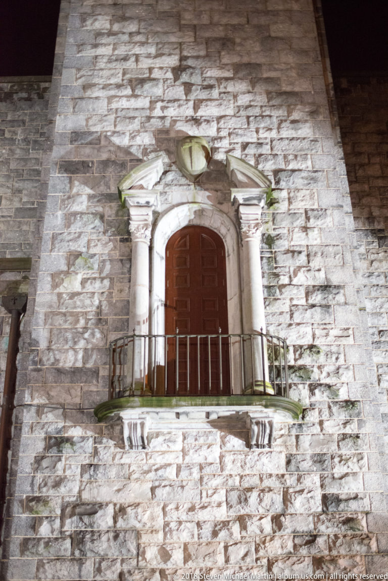 Elevated wooden doorway with ornate pale stone surround and small metal-railed balcony on rough cathedral masonry.