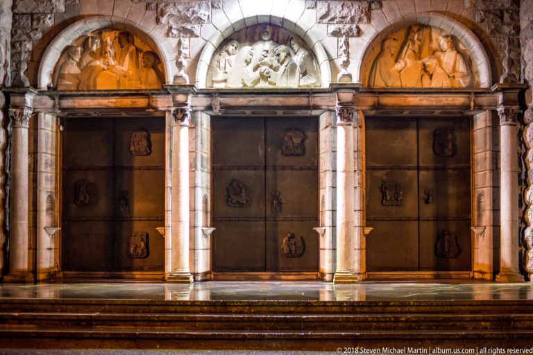 Three dark bronze cathedral doors with relief panels between pale columns and sculpted arches, reached by stone steps.