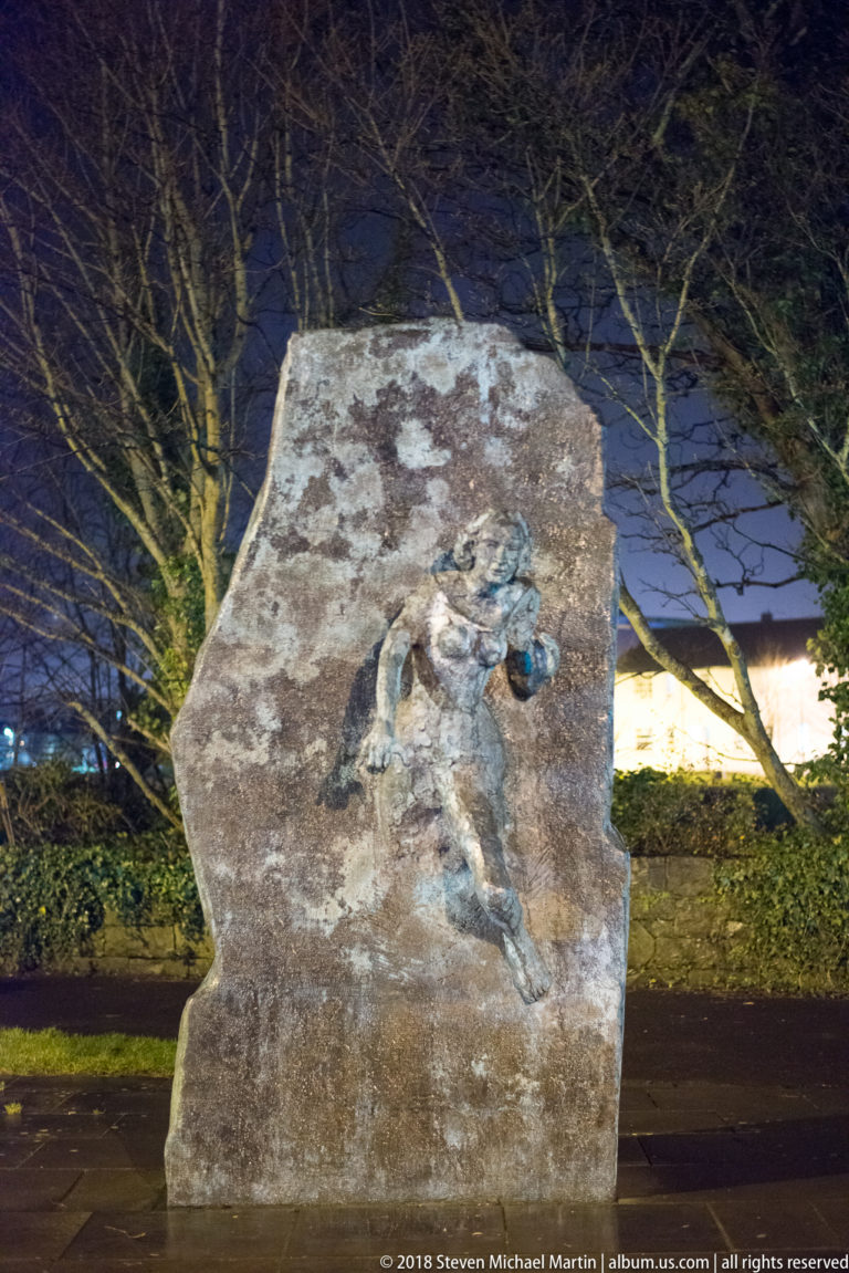 Rough upright stone memorial with a small bronze relief figure, set outdoors among trees at night.